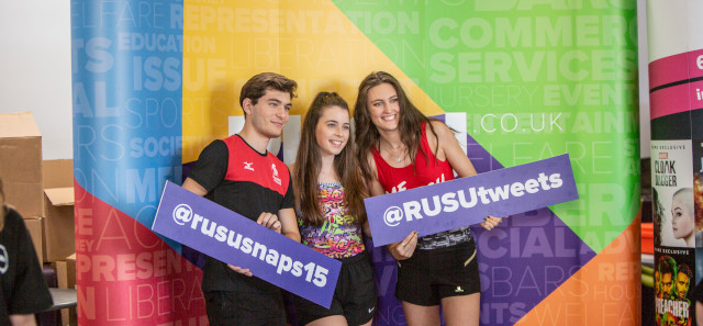 三名学生站在彩色背景前，手持两张纸板，宣传拉苏大学的社交媒体。一个写着“@rususnaps15”，另一个写着“@RUSU tweets”。