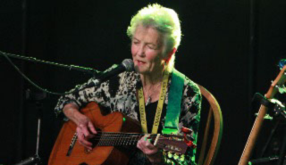 佩吉·西格(Peggy Seeger)坐在黑色背景下，弹着吉他，唱着歌