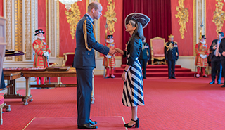 威尔士亲王殿下向副校长Parveen Yaqoob教授颁发大英帝国勋章。王子穿着军装，雅库布教授穿着深色和浅色条纹的连衣裙，深色夹克，戴着帽子。他们站在一个铺着红色地毯和金色装饰的房间里。